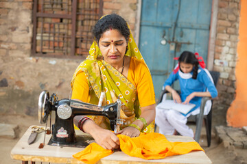 Rural indian woman using sewing machine while her young daughter studying behind her.