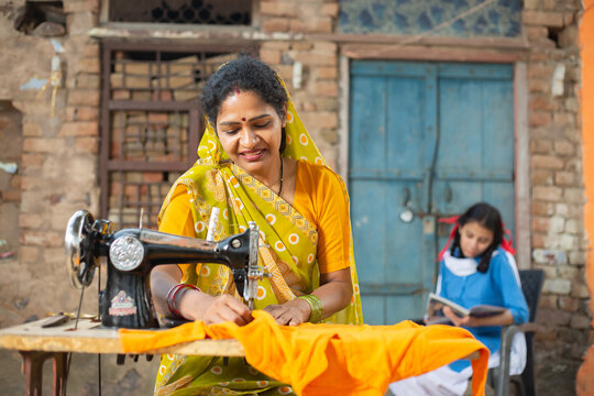 Rural Indian Woman Using Sewing Machine While Her Young Daughter Studying Behind Her.