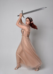  Full length portrait of pretty female model with red hair wearing glamorous fantasy tulle gown, crown. Holding a sword weapon, on studio background.