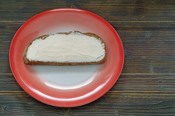 One slice of bread spread with soft white cheese on red plate. Dark rustic table, copy space