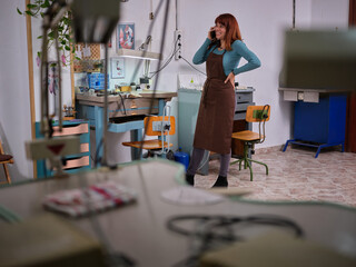 A female worker with apron standing in her jewelry artisan workshop while talking with her mobile phone