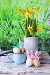 Easter eggs, metal vintage jug with a bouquet of fresh blooming yellow daffodils on a wooden table against the background of green grass, congratulations concept, village, farm