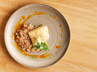 Fish lunch dinner at the restaurant. Fish fillet is served with rice, side dish, pearl barley, orange sauce and herbs in a gray plate. The concept of dietary nutrition. Top view on wood background.