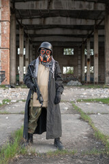 A soldier of the future after the nuclear apocalypse among the ruins: wearing a helmet, steampunk goggles, an orange respirator, a raincoat and camouflage. He holds a machine gun in his hands.