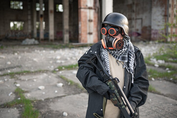 A soldier of the future after the nuclear apocalypse among the ruins: wearing a helmet, steampunk goggles, an orange respirator, a raincoat and camouflage. He holds a machine gun in his hands.
