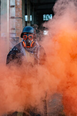 Obraz premium A soldier after a nuclear apocalypse among the ruins: wearing a helmet, steampunk glasses, a gas mask, and a raincoat. In his hands he holds a machine gun, a red smoke screen around him