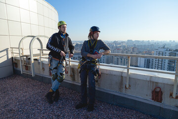 Two industrial climbers are standing on the edge of the roof leaning on the handrail