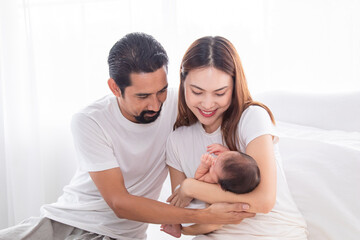 Fototapeta premium Selective focus of Asian man and woman holding newborn baby in bed at home. Happy family smiling with adorable infant, young parents holding little sweet toddler baby, mom hold toddler with love.