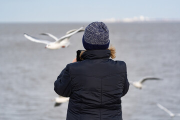 Eine junge Frau mit Mütze am Strand fotografiert fliegende Möwen mit dem Handy