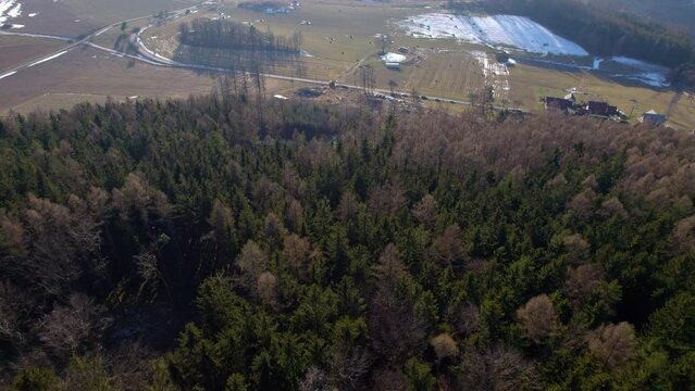 Hill with dense forests above the village with houses, fields, straw bales and parked tourists - view down from the drone