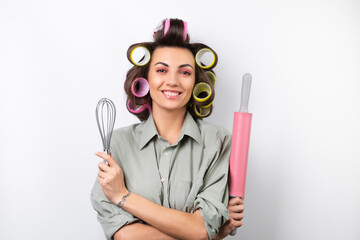 Beautiful housewife. Young cheerful woman with hair curlers, bright make-up, a rolling pin and a whisk in her hands, on a white background.