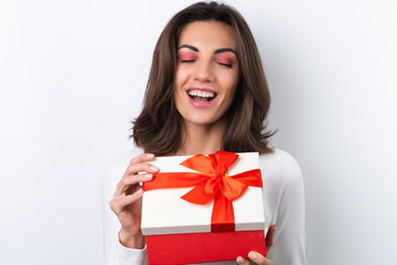 Fototapeta premium Young woman in a cocktail dress, gold chain, bright spring pink makeup on a white background. Holds a gift box for March 8 and smiles cheerfully.