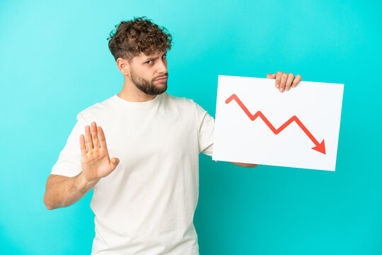 Young handsome caucasian man isolated on blue background holding a sign with a decreasing statistics arrow symbol and doing stop sign