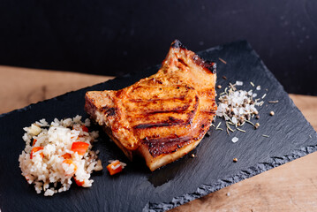 Pork steak on a rib lies on a slate board, next to rice with vegetables, coarse salt with spices