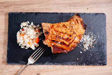 Pork steak on a rib lies on a slate board, next to rice with vegetables, coarse salt with spices