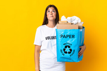 Young brazilian girl holding a recycling bag full of paper to recycle isolated on yellow background and looking up