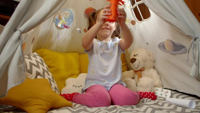Little girl is playing an alien from outer space with a paper humanoid sitting in a tent at home. Child lets the alien between the planets smiling cheerfully.