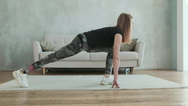 Young attractive woman with prosthetic leg does stretching exercise for muscles of body inside house. concept of sports and fitness for disabled. Yoga for health. Active lifestyle of disabled people