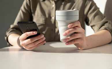 Woman hands holding phone and eco cup. Closeup. Woman sitting at table and using smartphone for typing message, surfing Internet, paying online. High quality photo