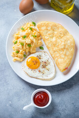 White plate with scramble, omelette and fried eggs, elevated view on a light-blue stone surface, vertical shot