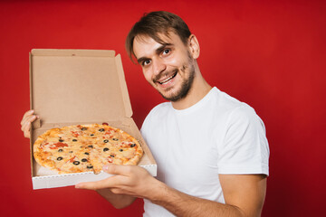 A man with a smile in a white t-shirt on a red background holds a box with an appetizing pizza in his hands. Pizza delivery man