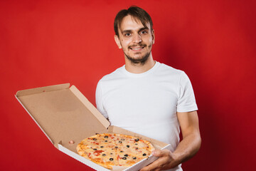A man with a smile in a white t-shirt on a red background holds a box with an appetizing pizza in his hands. Pizza delivery man