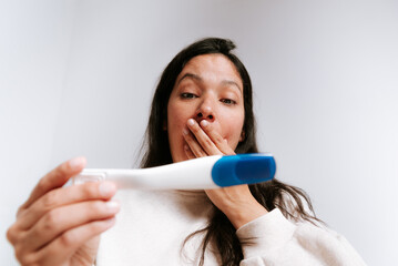 young adult woman looking at pregnancy test with happy expression in her bedroom. latina woman with dark hair in her thirties smiling at home discovering she is pregnant and will be a single mom