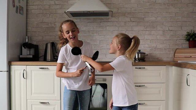 two cute blonde sister girls dance in a modern white kitchen, sing in spoons, dance, rejoice and have fun at home, great mood, songs and fun