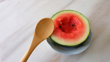 Small watermelon cut in half, with a small hole in the middle, and a wooden spoon beside it. On a marble surface.