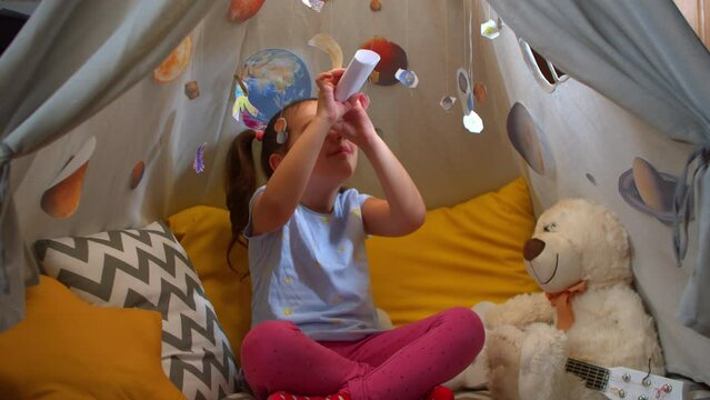 Little girl fantasizes about watching the planets with a homemade telescope and carved planets hanging in a tent in the simeiny interior. Preschool girl looks at the stars at night through a paper.
