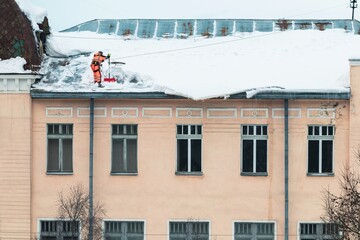 A working man in bright overalls with a safety belt with a shovel clears snow from the roof of an old building. Prevention of snow falling from the roof, industrial mountaineering