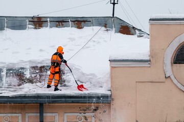 A working man in bright overalls with a safety belt with a shovel clears snow from the roof of an...