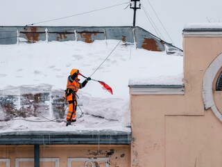 A working man in bright overalls with a safety belt with a shovel clears snow from the roof of an old building. Prevention of snow falling from the roof, industrial mountaineering