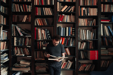 Adult student reading a book in the college library. Young standing male wearing glasses with...