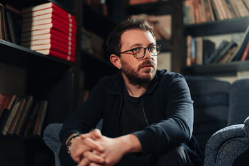 Positive male student wearing glasses in library with bookshelves on background. Proud and successful small business owner