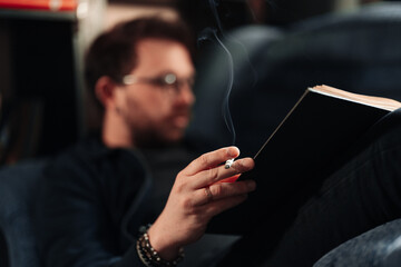 Man smoking and reading, holding a book and a cigarette in hands while lying on a couch. Focus on cigarette.