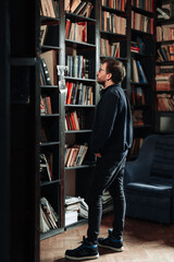 Adult student looking for a book in the college library. Young standing male wearing glasses look to bookshelves.