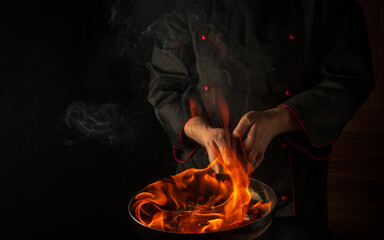 The chef cooking food in pan with fire flame on black background. Restaurant and hotel service...