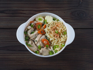 Chicken soup with chinese noodles, vegetables and meat in asian style in a white bowl on a dark wooden table, top view, flat layout. Hot broth with instant vermicelli. Traditional oriental cuisine