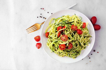 Zucchini pasta topped with tomatoes and basil pesto. Top view on a white marble background. Healthy eating, low carb diet concept.