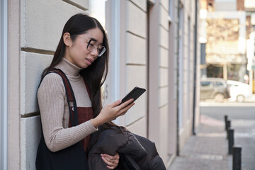 Portrait of a young asian woman checking the phone wearing glasses and overall skirt standing up...