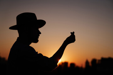Silhouette of man in hat at sunset showing Korean heart sign using hands. Hipster male showing 'mini heart' gesture with urban view. Love or romance concept. High quality image