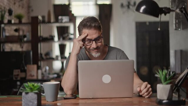 Bearded man working online with laptop computer at home sitting at desk. Home office, browsing internet. Portrait of mature age, middle age, mid adult man in 50s.