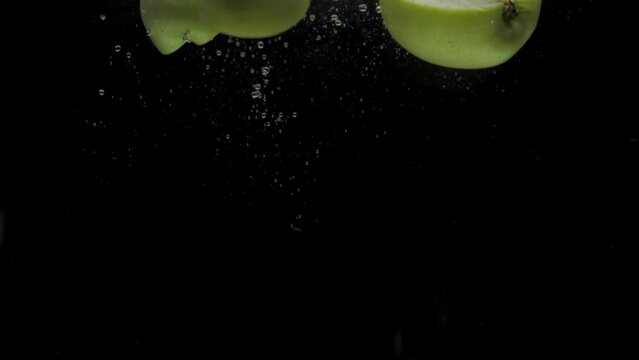 Slow motion green apple halves falling into transparent water on black background. Fresh fruit splashing in aquarium. Organic fruits, healthy food, diet, air bubbles