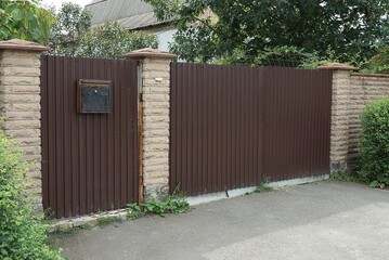 Fototapeta premium one big closed brown metal gate and part of the wall of the fence made of bricks in green vegetation on the street