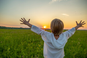 Happy boy with raised hands looks at the sunset  in summer. Boy in a field with his hands raised...