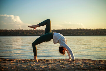 Young woman on sandy beach by the river doing fitness and yoga exercise on background of blue sky and white clouds