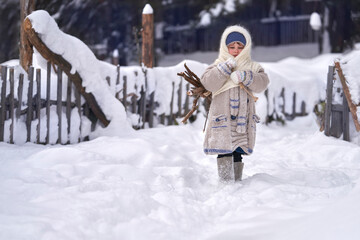 Rustic winter motif. Vintage. A little girl returns to the village with a bundle of collected firewood. She walks down a snowy street. Copy space.