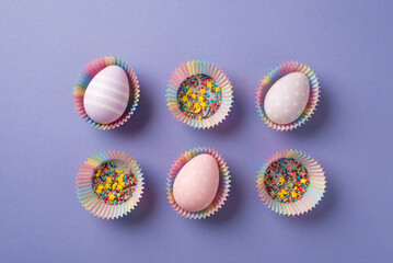 Top view of six multicolored cupcake cups with three pastel eggs and a lot of bright sprinkles inside on the isolated purple background copyspace