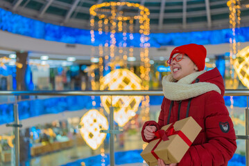 Fototapeta premium teenage girl in a white jacket in a mall holding a box with a gift on the background of a shop window with discounts during a sale